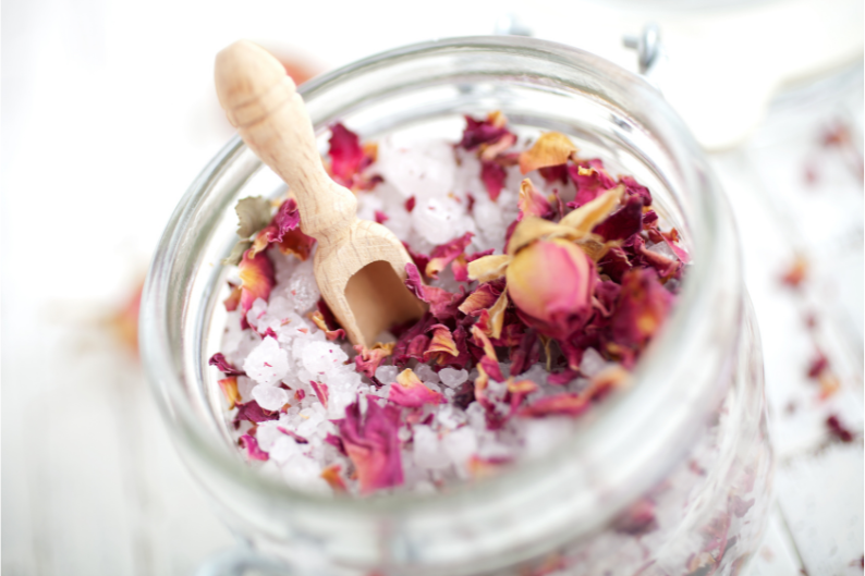 A jar filled with sea salt and rose petals with a wooden spoon.