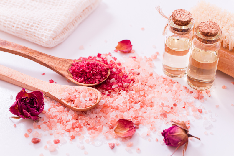 A wooden spoon filled with pink salt next to a bottle of essential oil.