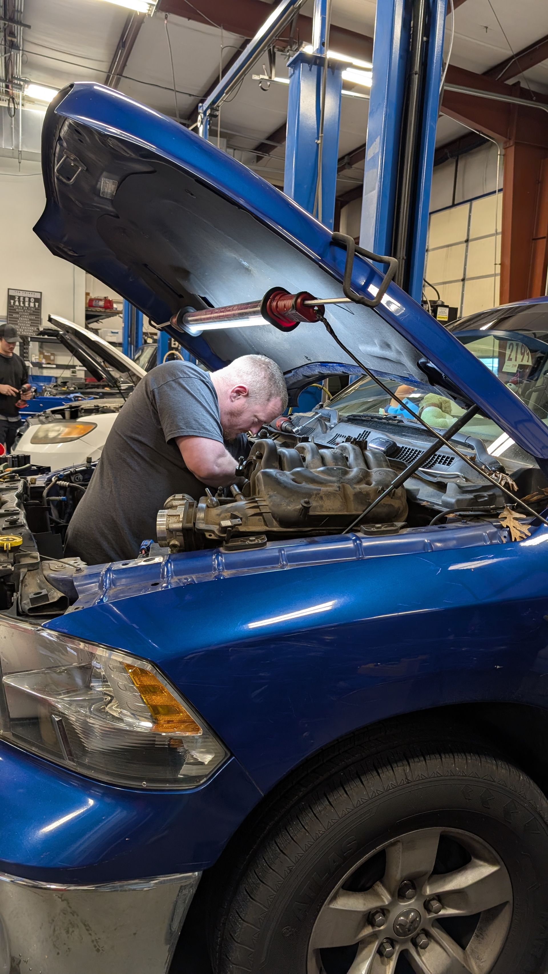 A mechanic works on the engine of a blue pickup truck inside an auto repair shop.