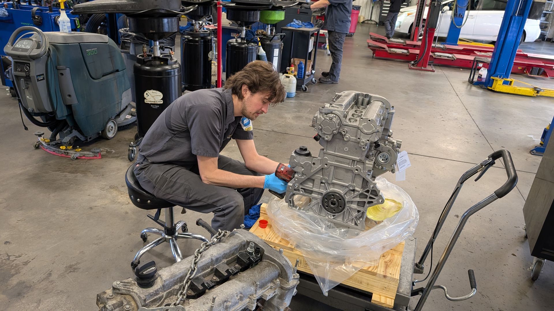 A technician works on an engine block placed on a wooden pallet inside an automotive repair shop.