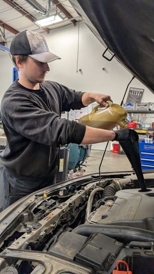 A mechanic in a cap and dark sweater pours oil into an open car engine in a garage.