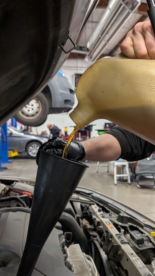 A mechanic wearing black gloves pours golden oil into a car engine using a black funnel in a service garage.
