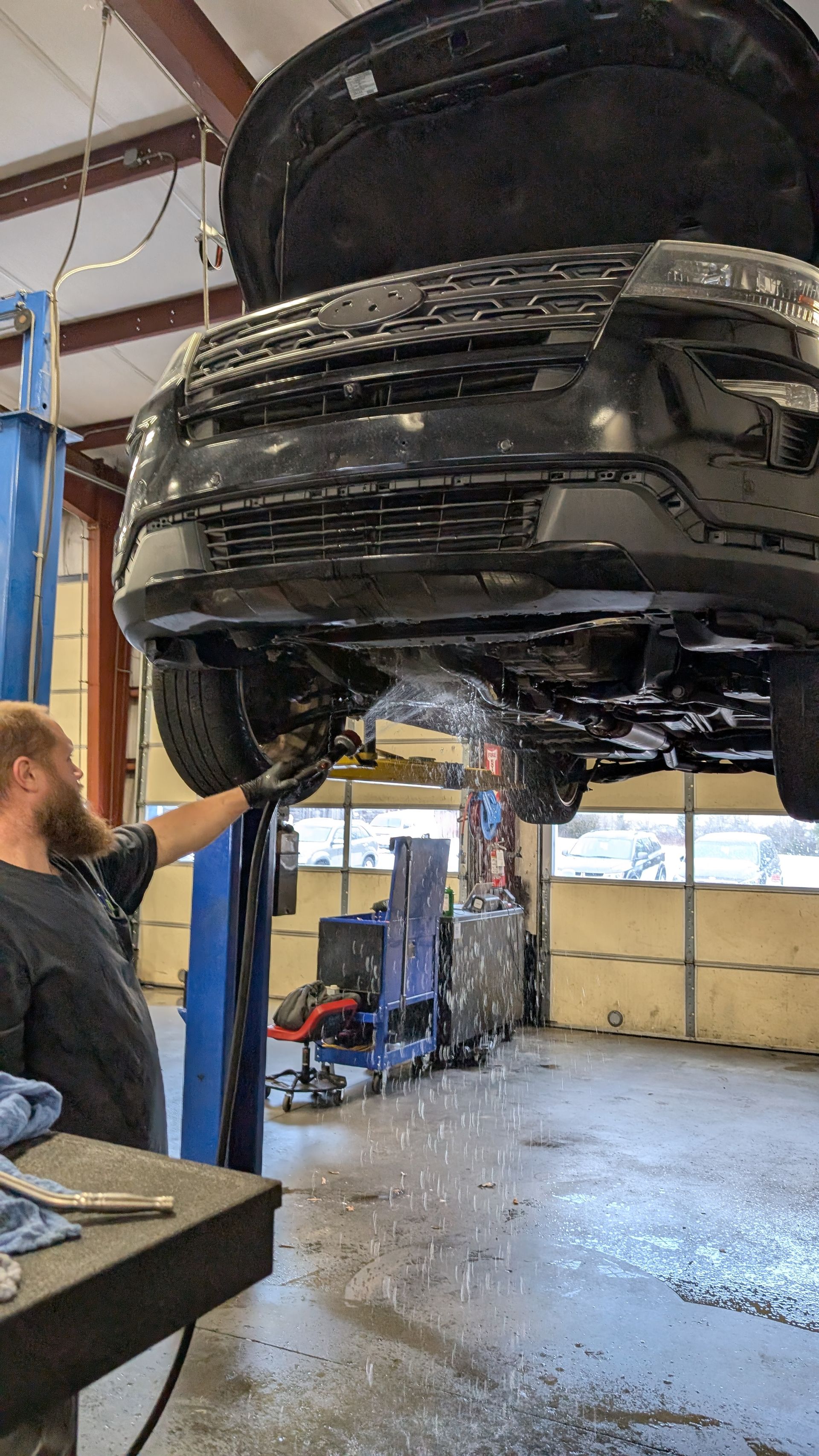 A mechanic works under a black car raised on a lift in a garage as fluid drains from the engine bay.