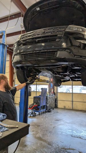 A mechanic in an auto repair shop works under a black car hoisted on a lift as fluid drains from the engine.