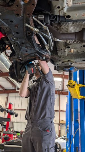 A technician in a gray uniform uses a power tool to repair the undercarriage of a vehicle raised on a shop lift.