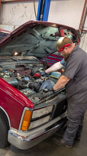 A mechanic in a grey uniform and red safety glasses repairs a red pickup truck with the hood open inside a workshop.