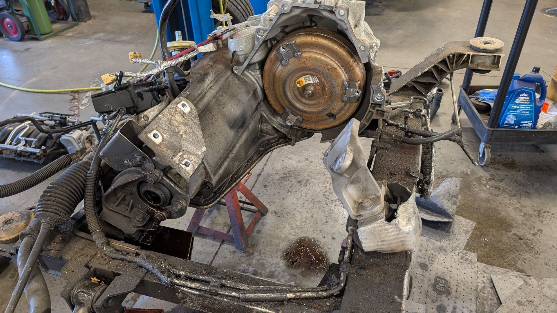 A disassembled automotive transmission with a prominent torque converter, resting on a stand in a garage setting.