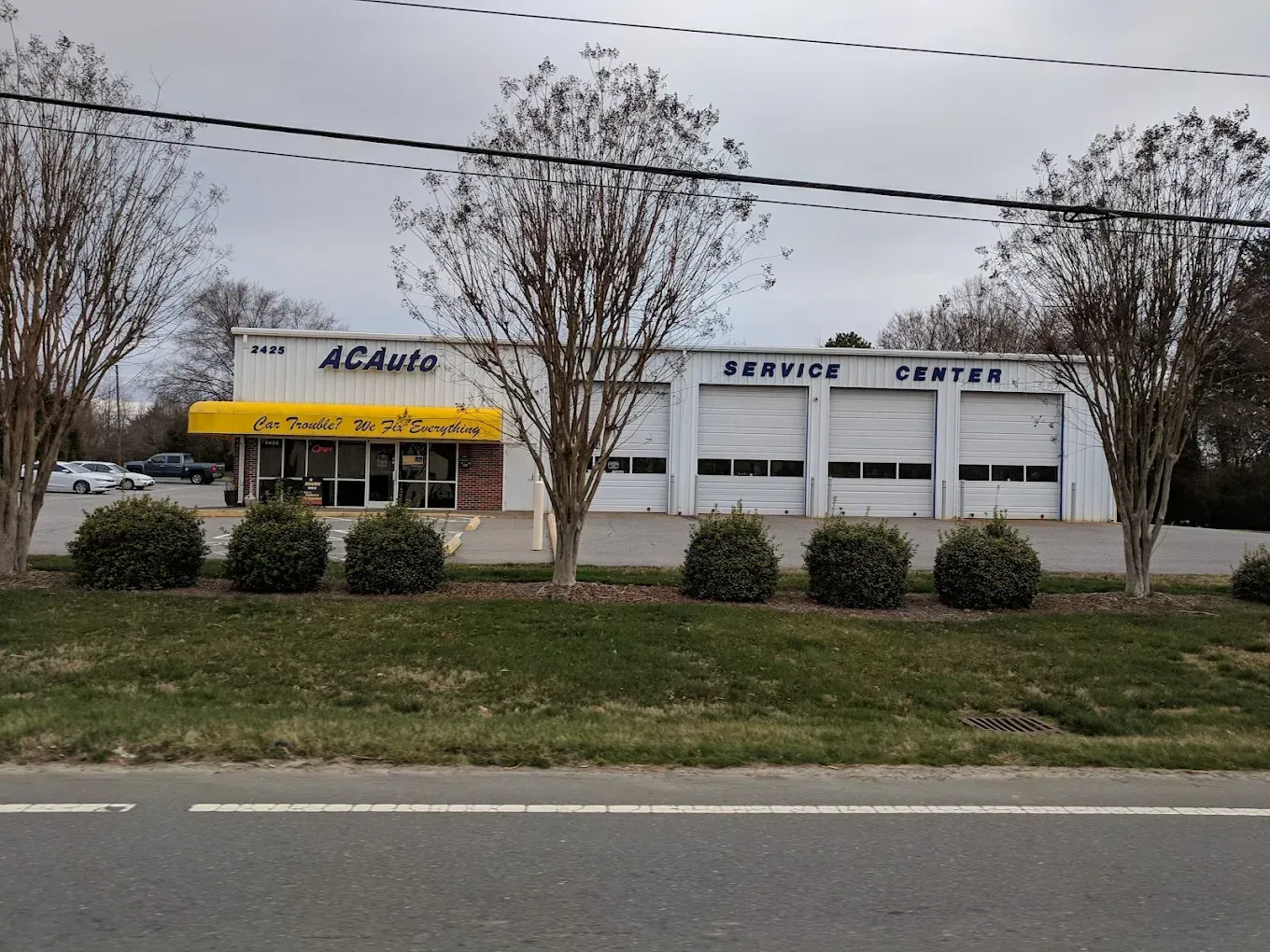 A white one-story AC Auto Service Center building with a yellow awning, three garage bays, and trees in the foreground.