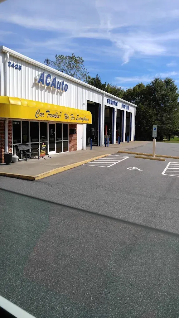The exterior of an ACAuto repair shop with a yellow awning and white siding on a sunny day with a parking lot in front.