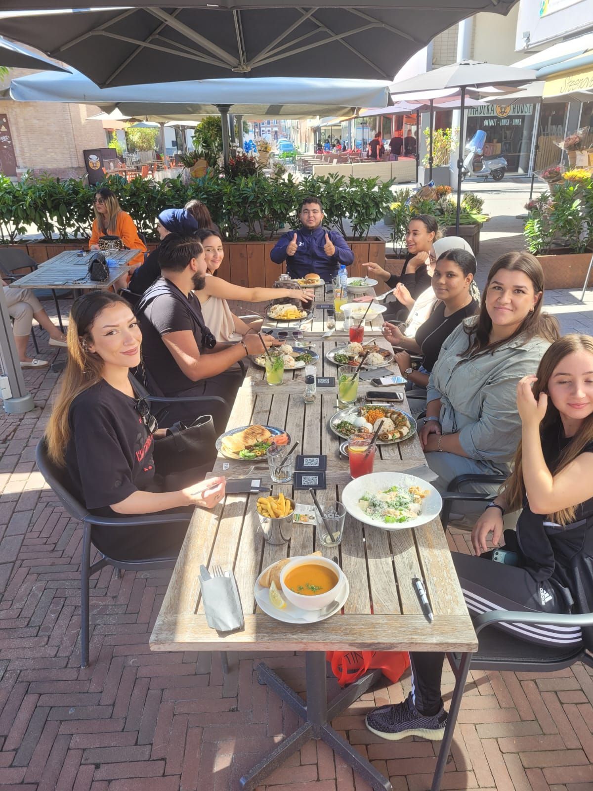 Mensen dineren in een restaurant in de openlucht, zittend rond een lange houten tafel met borden en drankjes onder parasols.