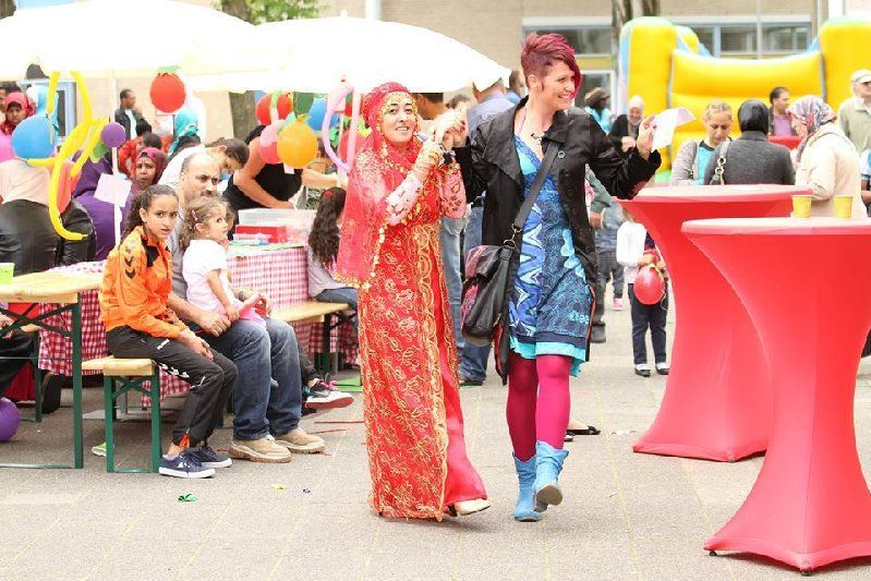 Twee artiesten in kleurrijke kostuums lopen door een openluchtevenemententerrein met tafels en zittende toeschouwers.