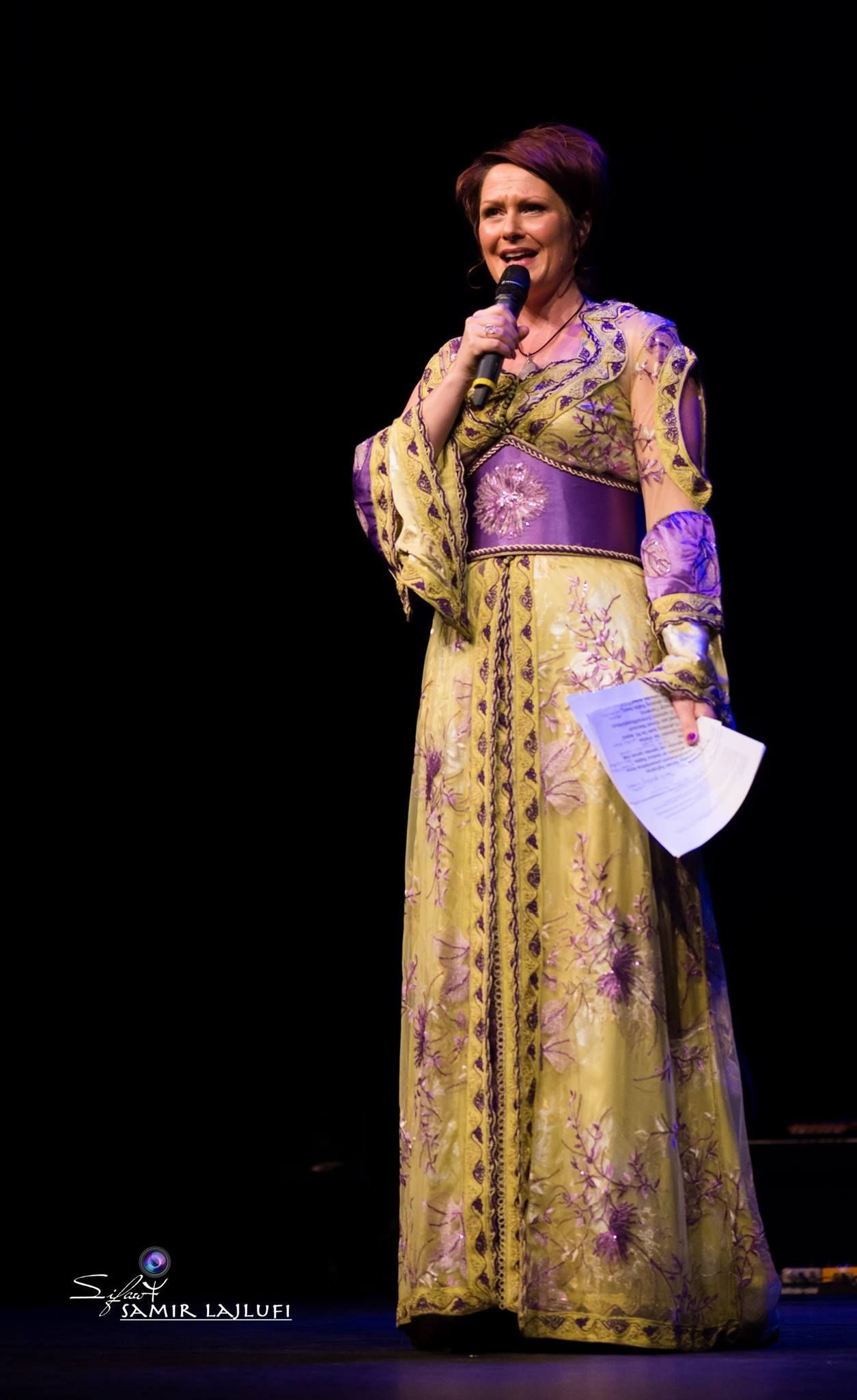 Vrouw in een gele jurk met patroon, met een microfoon en papier in haar handen, op een donker podium.