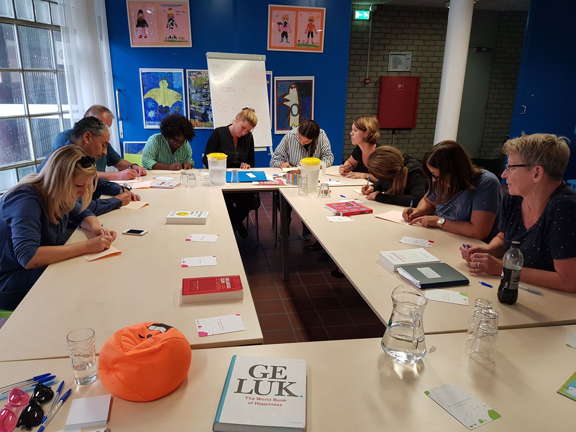 Mensen zitten rond een vergadertafel in een kleurrijke vergaderruimte, te overleggen en te schrijven.