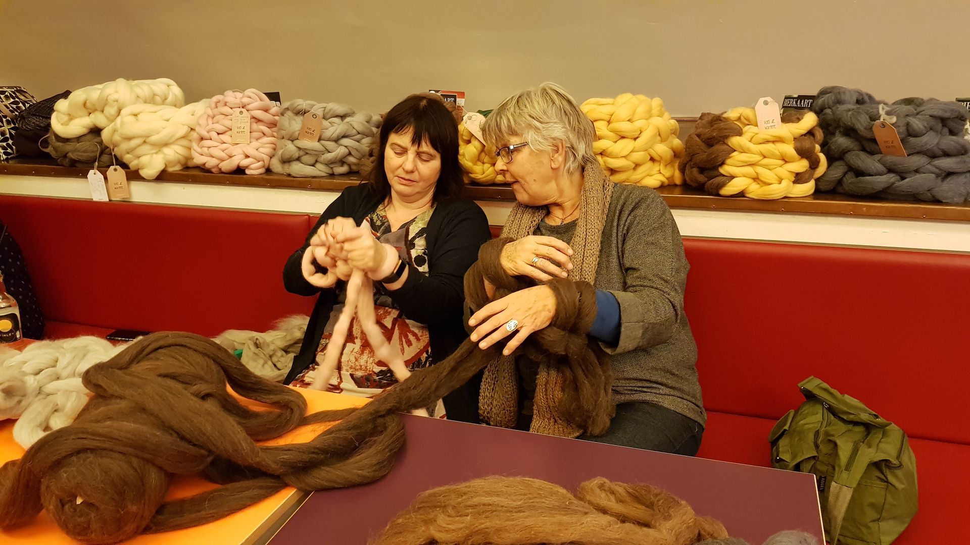 Twee vrouwen bekijken garen in een winkel, zittend aan een tafel met kleurrijke stapels wol achter hen.
