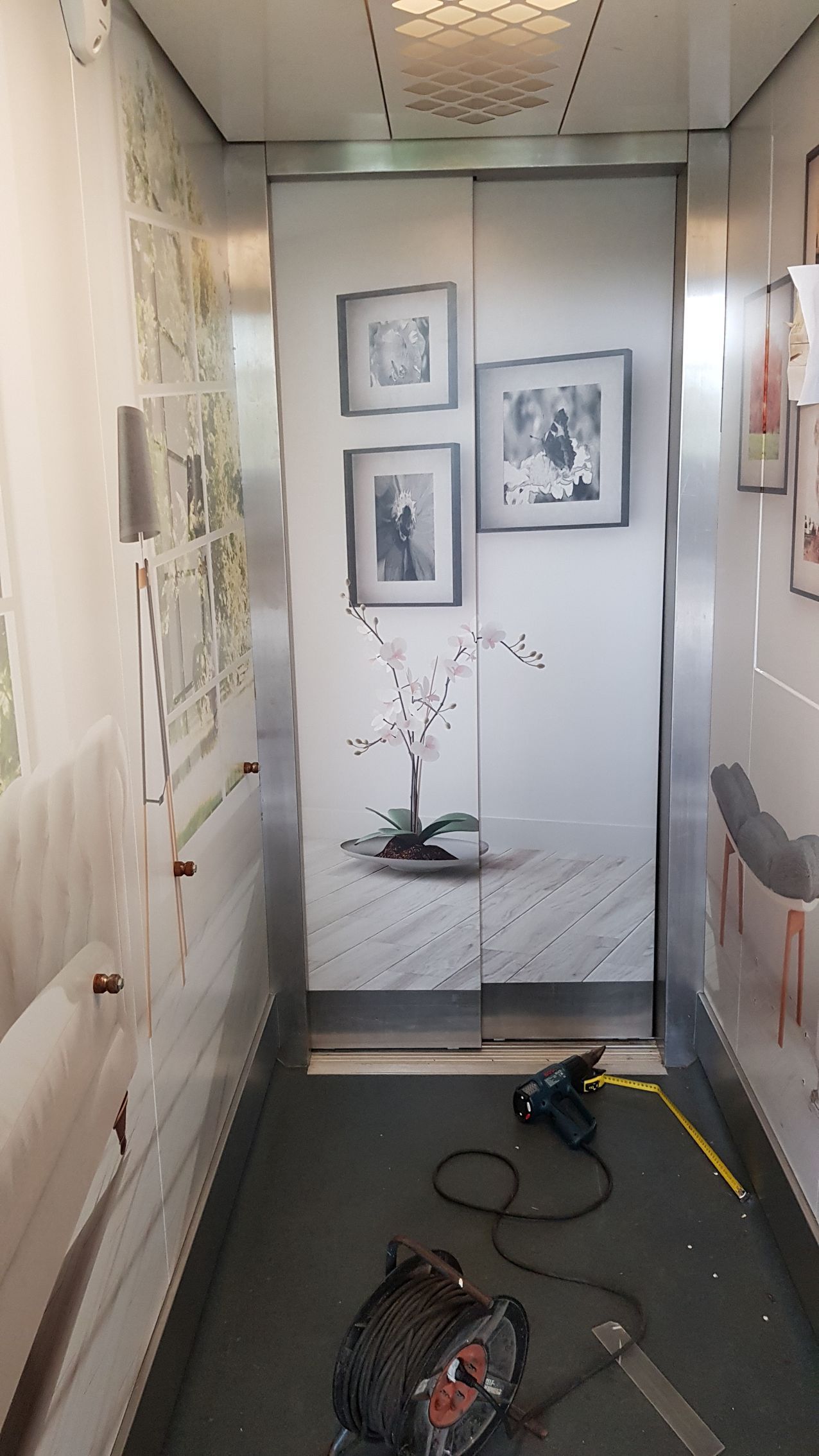 Smalle gang met ingelijste kunst aan de muren, een kleine orchidee op een tafel en gereedschap en kabels op de vloer.
