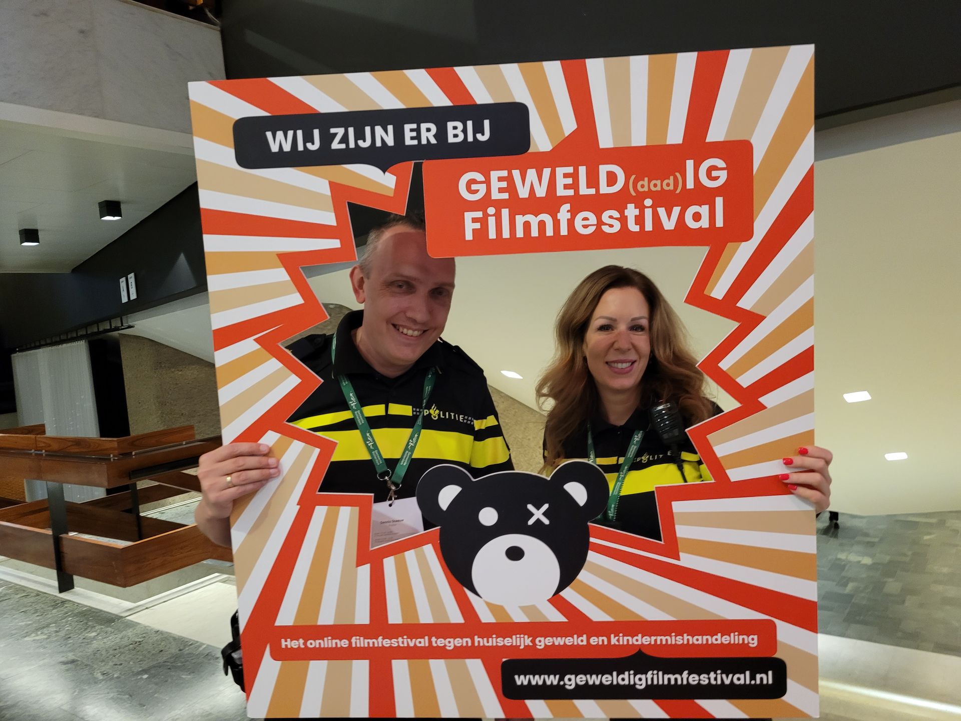 Twee mensen houden een rood-wit promotiebord vast voor het Gevelde Filmfestival in een overdekte zaal.