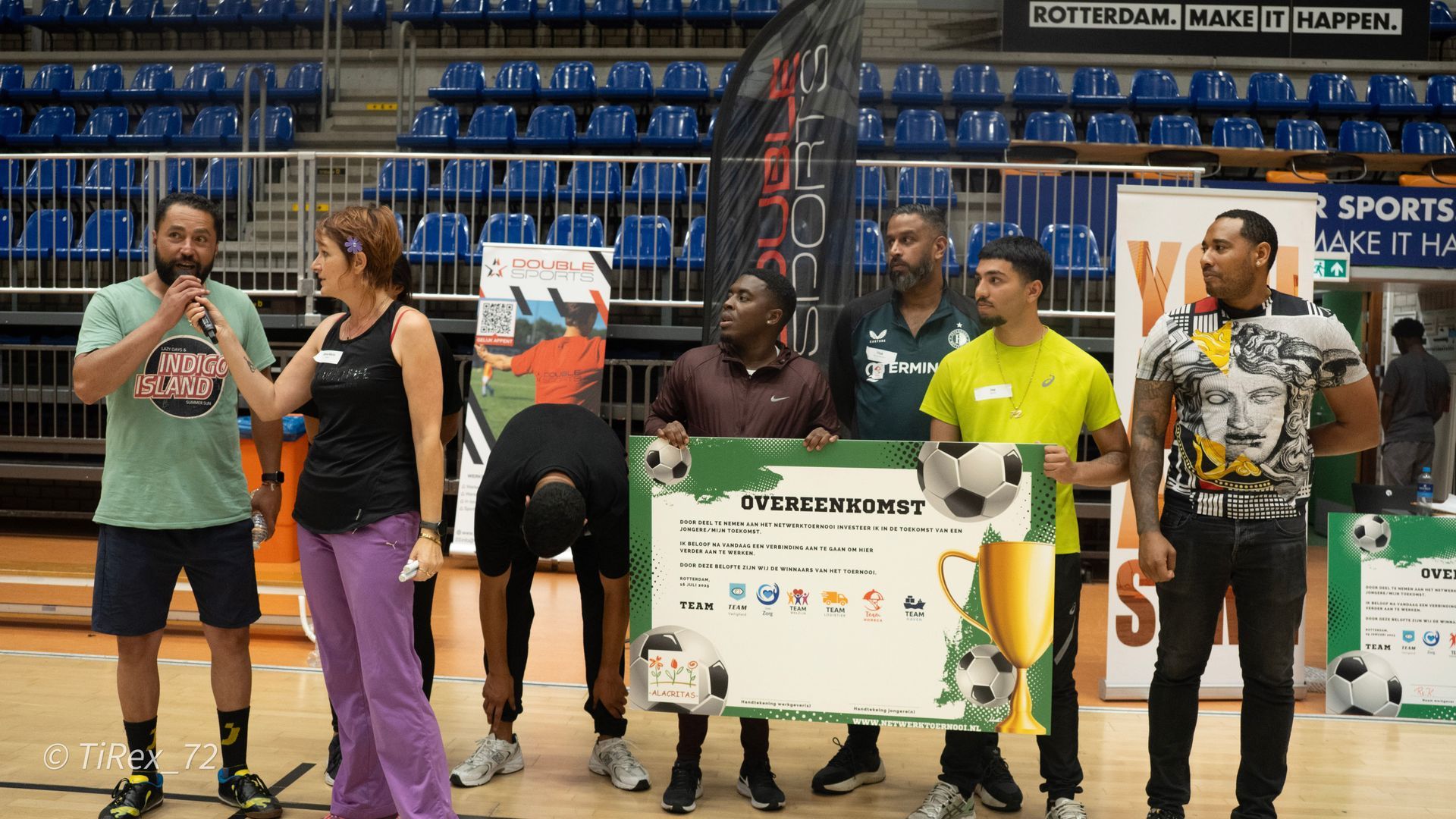 Mensen die een grote cheque vasthouden op een basketbalveld met blauwe tribunes op de achtergrond.