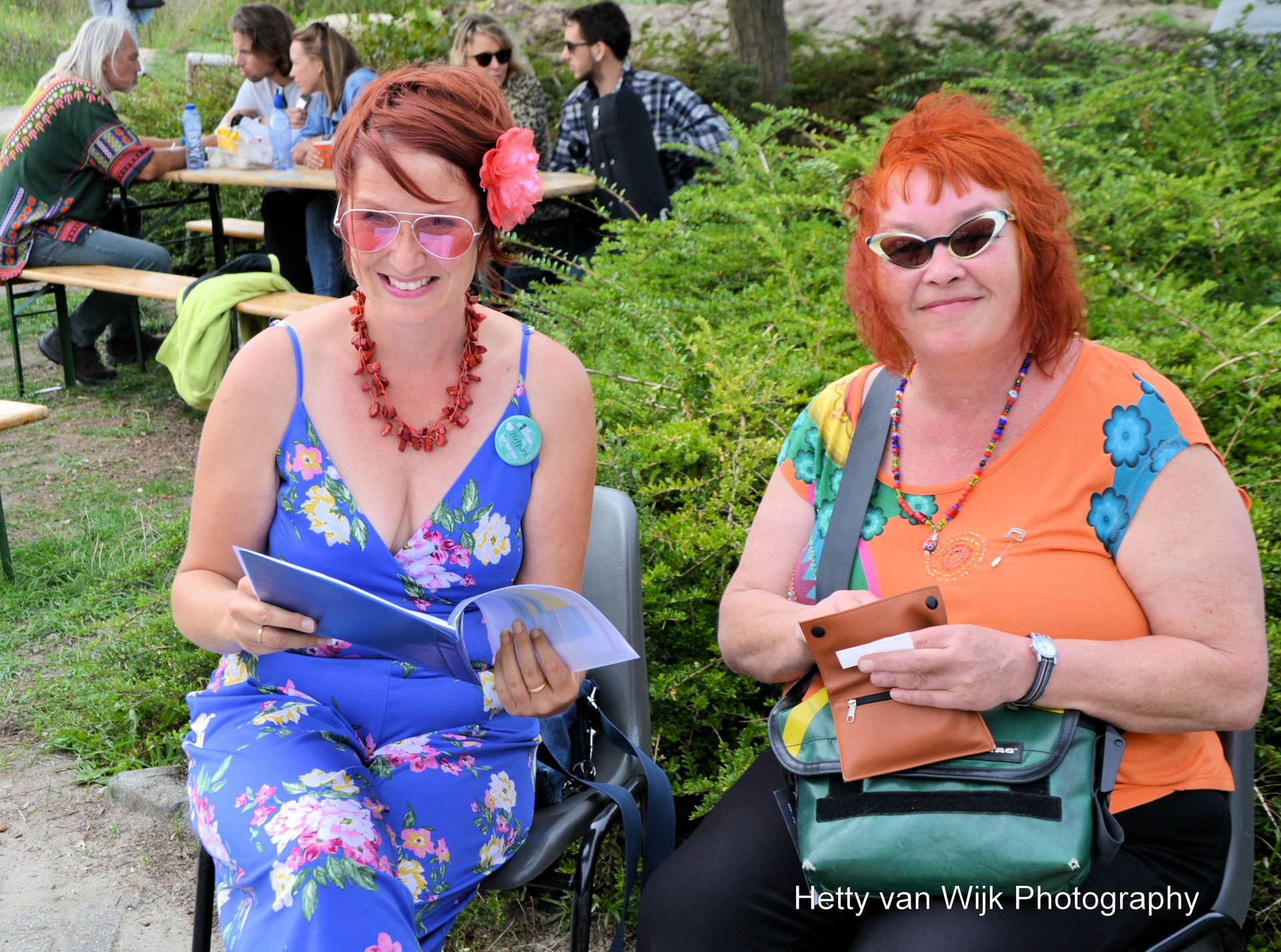 Twee mensen zitten lachend op een parkbankje, met papieren in hun handen, en op de achtergrond picknicktafels en groen.