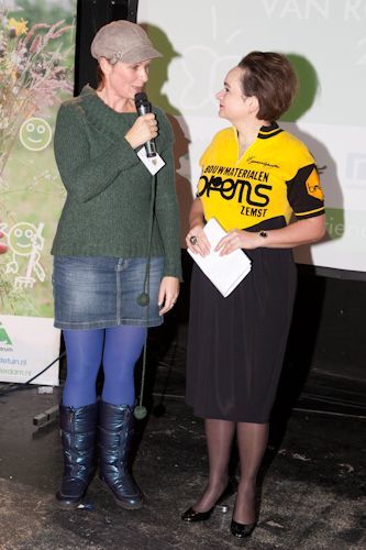 Twee vrouwen spreken op een podium, de ene met een microfoon in de hand en de andere met aantekeningen.