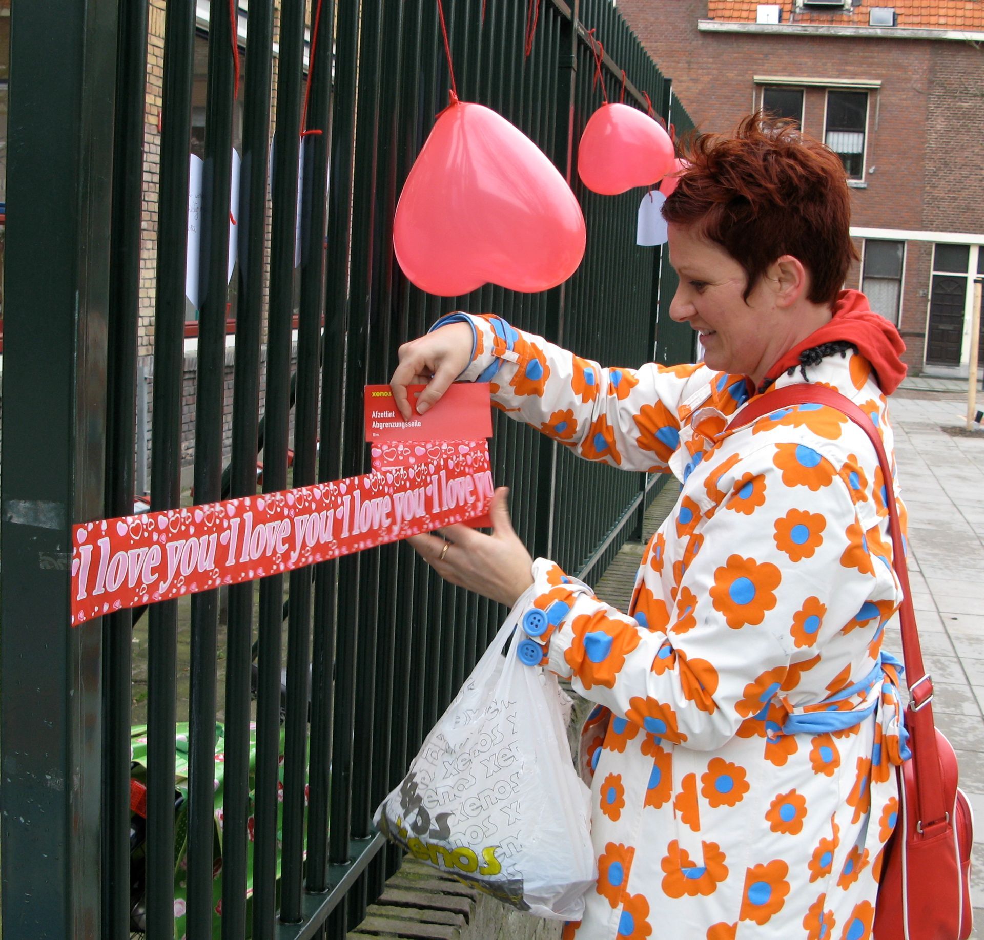 Een persoon in een jas met bloemenprint hangt een rood lint aan een hek, met roze ballonnen in de buurt.