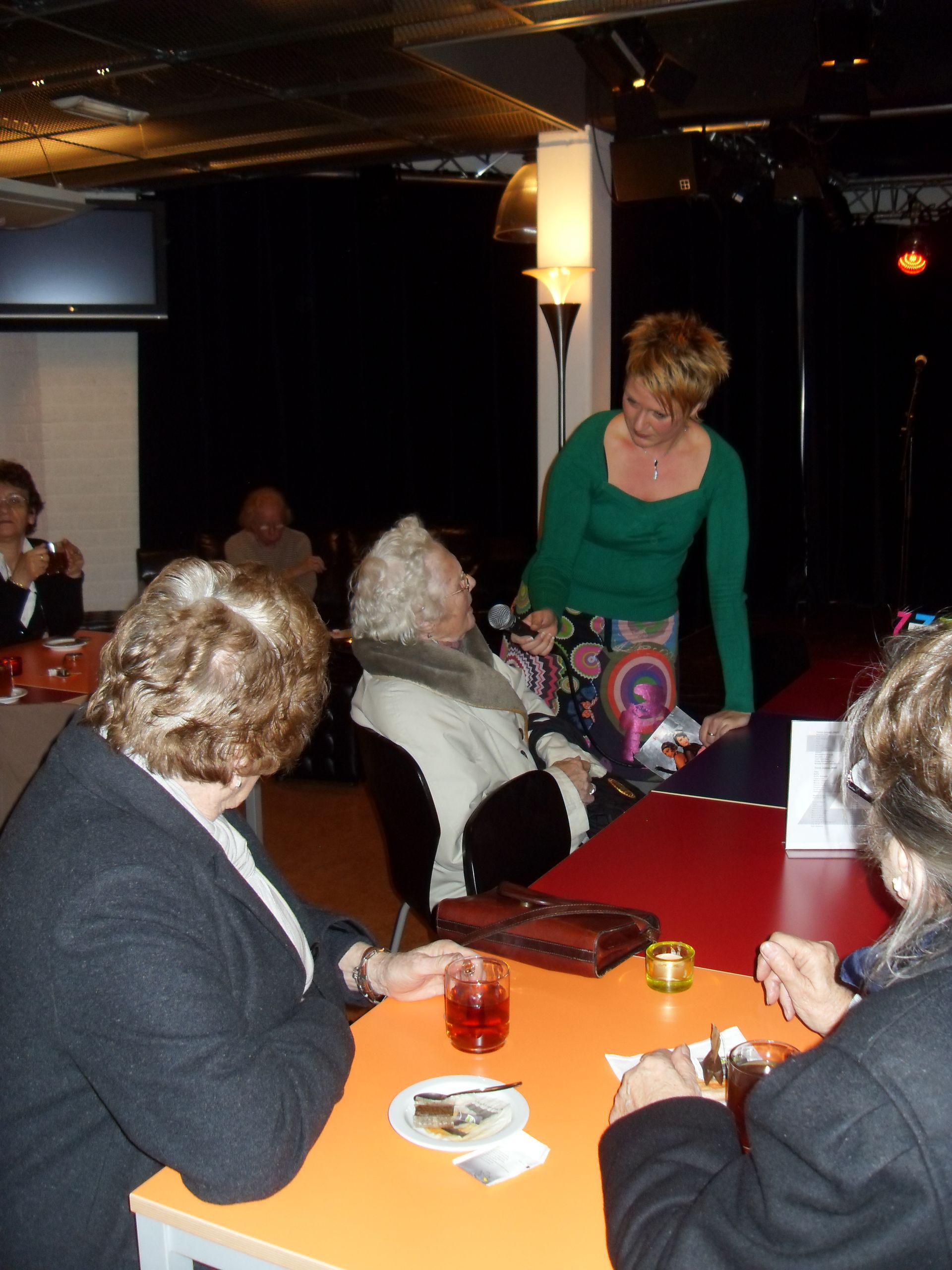 Mensen zitten aan een restauranttafel, terwijl een vrouw in een groene top vlakbij staat in een schemerige eetzaal.