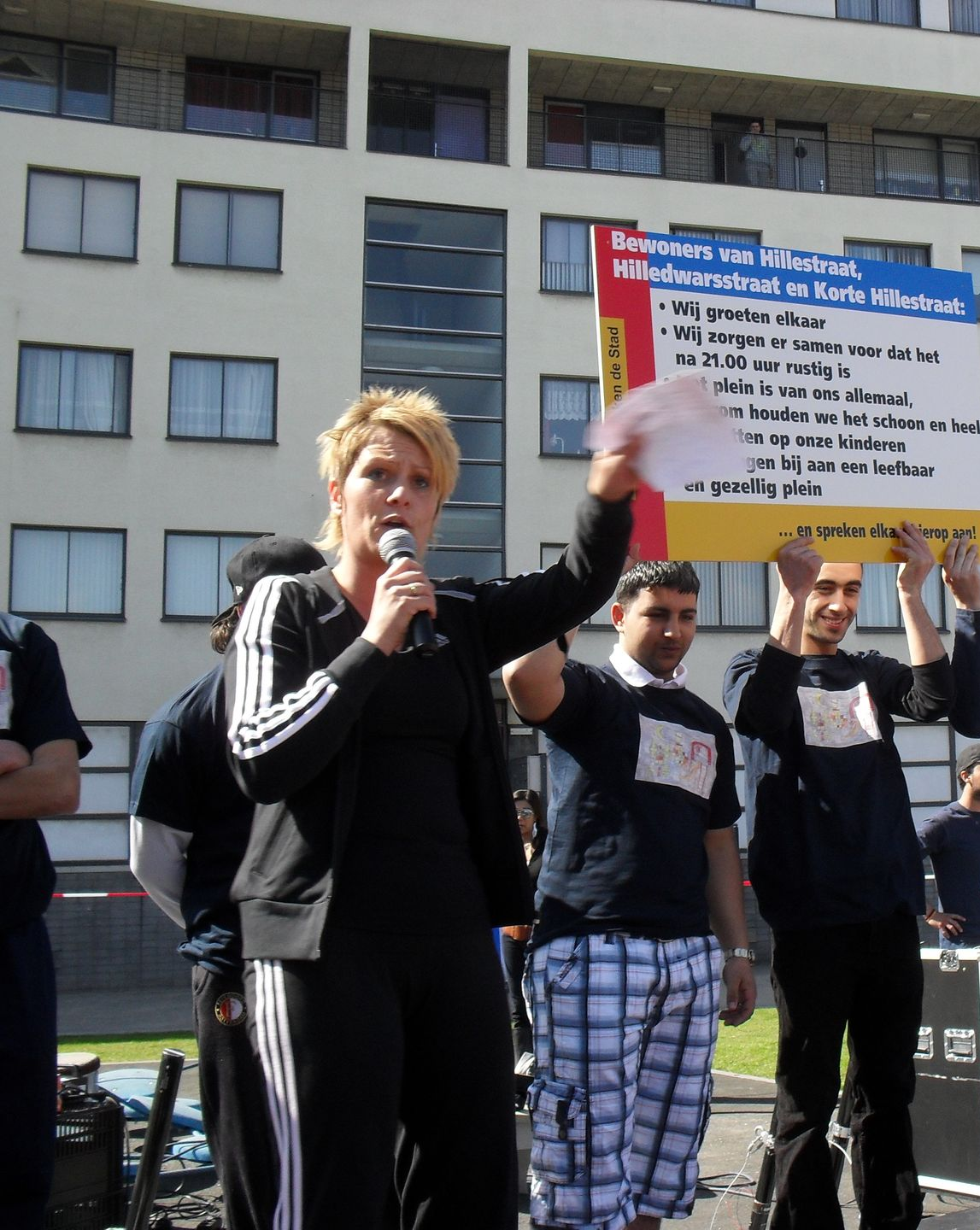 Een vrouw spreekt in een microfoon tijdens een protest en houdt een bord vast voor een gebouw.