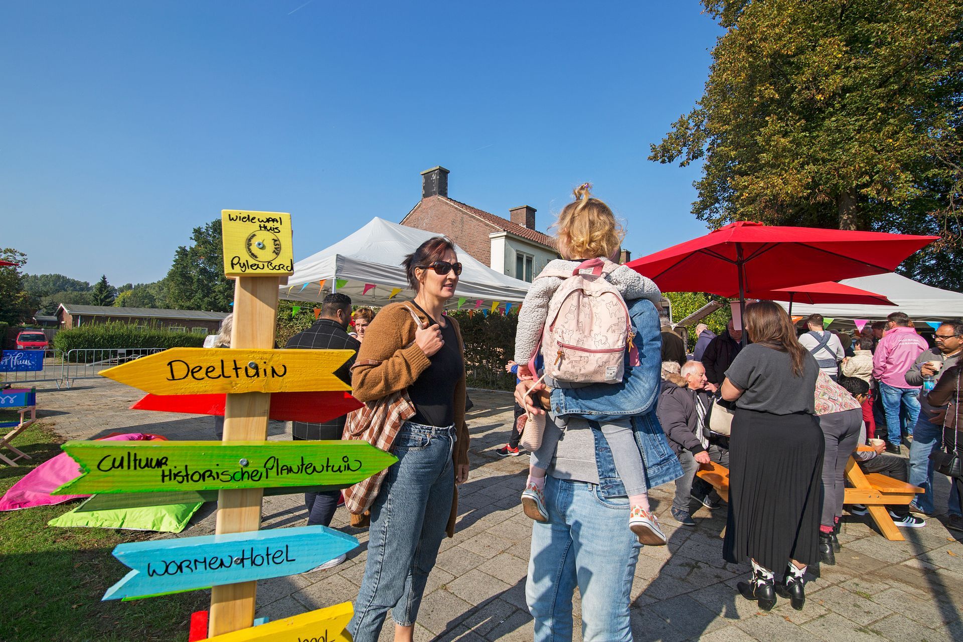Een kleurrijk uithangbord op een openluchtmarkt met mensen die op een zonnige dag langs rode parasols lopen.