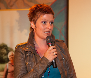 Vrouw met kort haar spreekt in een microfoon tijdens een evenement binnenshuis, gekleed in een bruine jas.