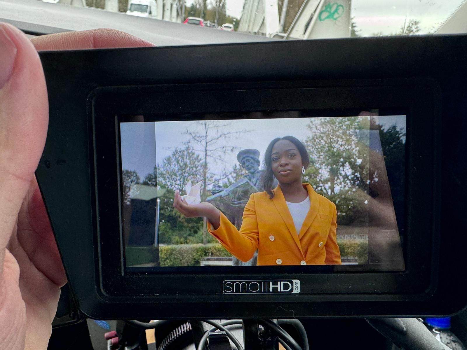 Op een scherm op het dashboard van een auto is een vrouw in een gele blazer te zien die buiten aan het praten is.