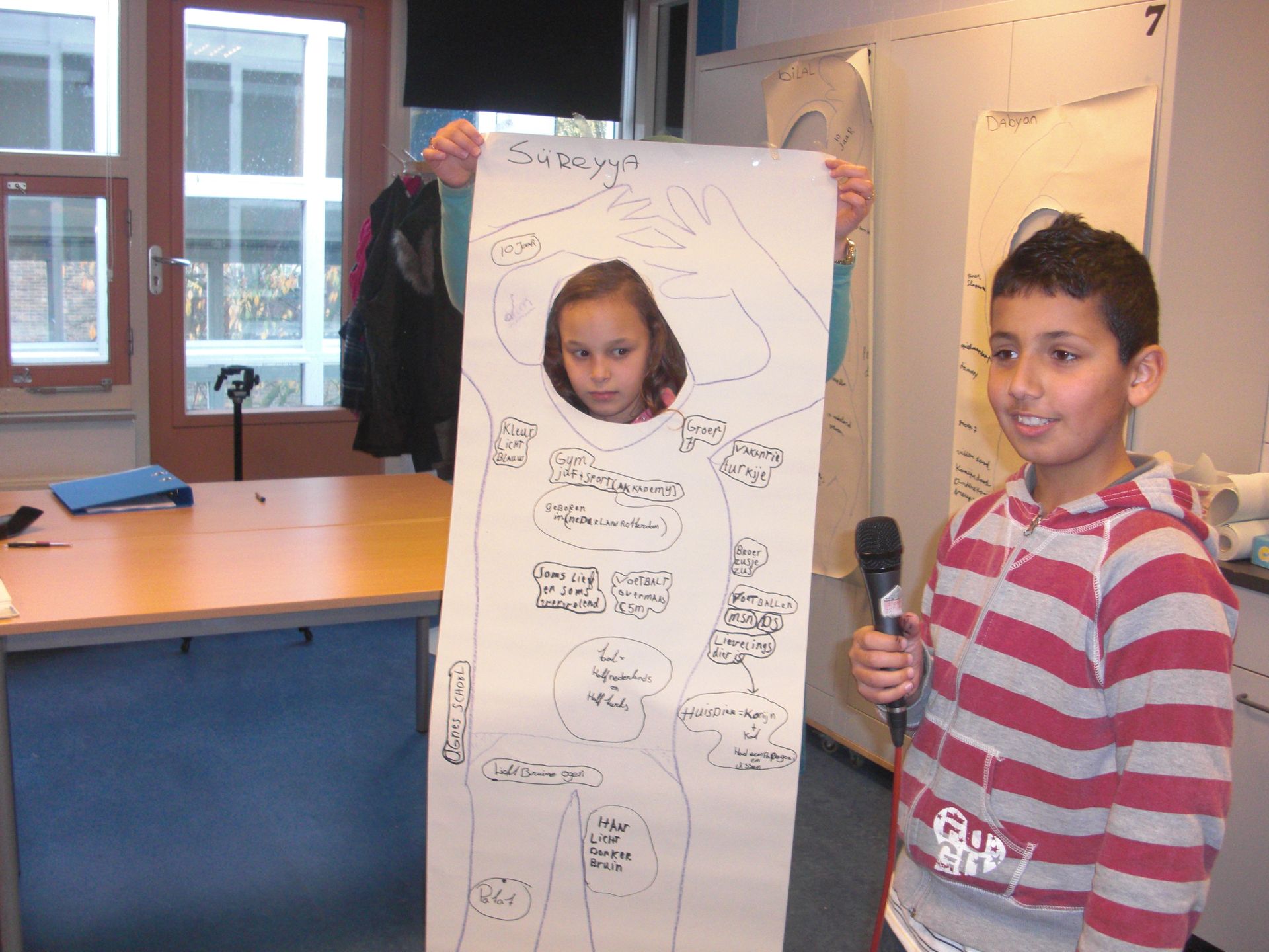 Twee kinderen in een klaslokaal houden een grote tekening op papier omhoog; een van hen lacht en houdt een microfoon vast.