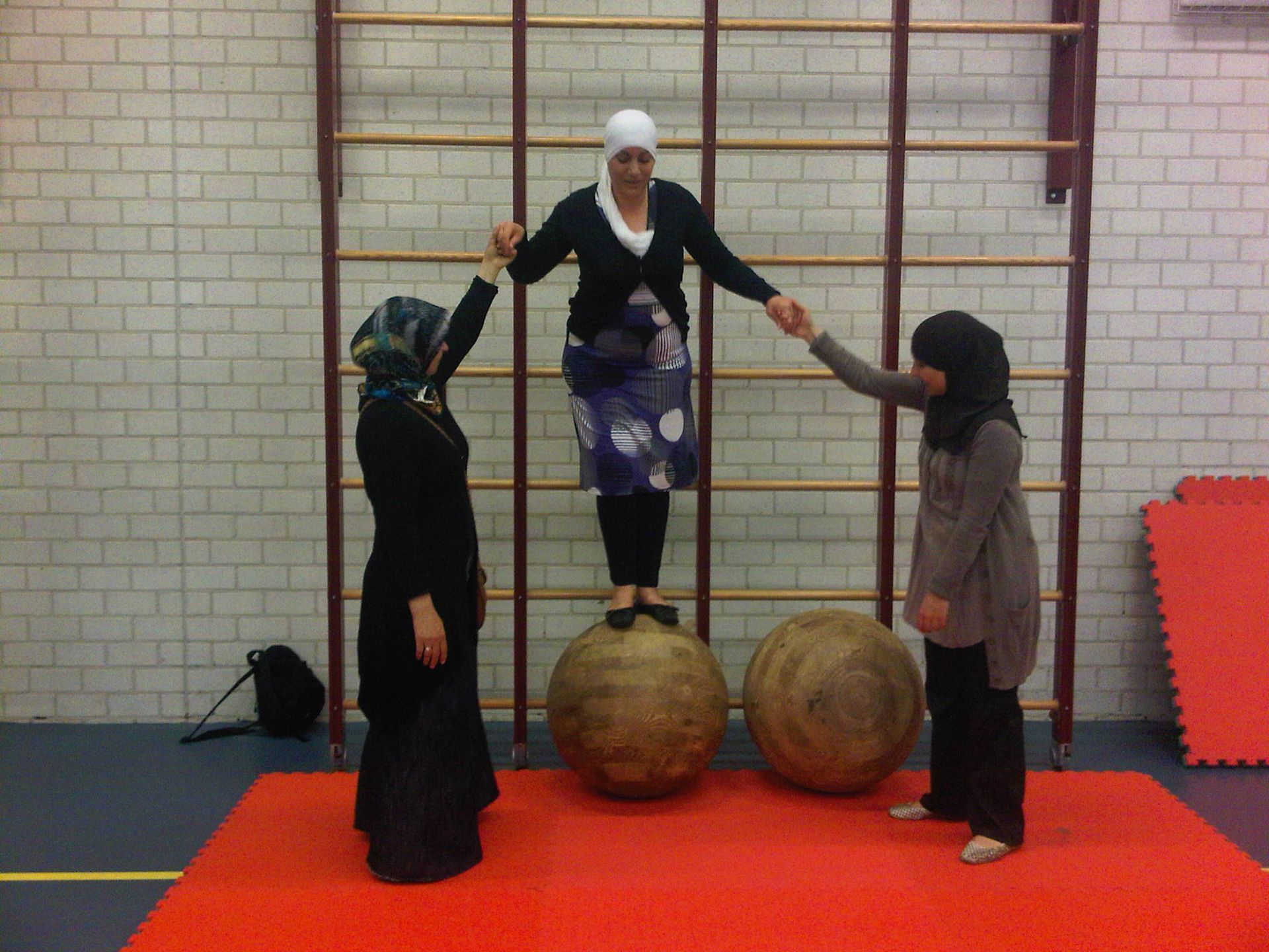 Drie mensen balanceren op fitnessballen in een sportschool en houden elkaars handen vast op een rode mat.