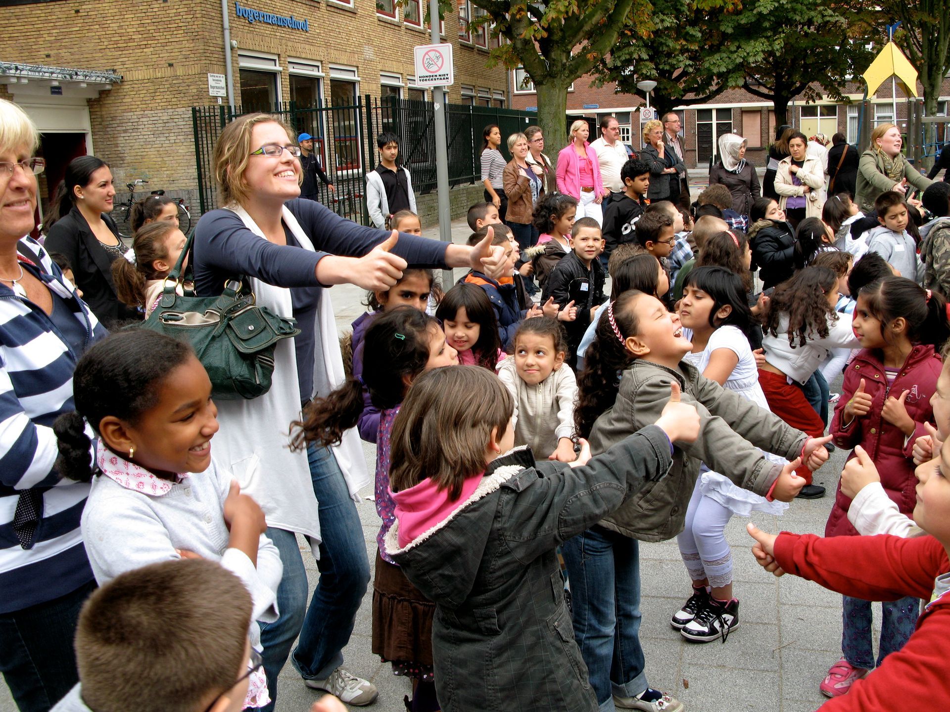 Kinderen dansen met volwassenen op een binnenplaats, hand in hand in een kring onder bomen en parasols.
