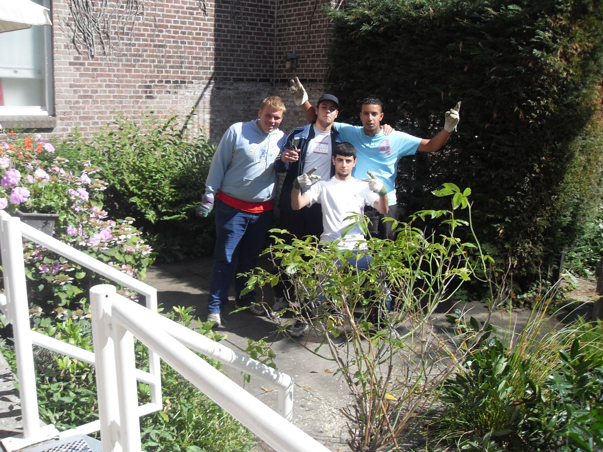 Vier mensen poseren buiten op een zonnig pad naast struiken en een bakstenen gebouw.