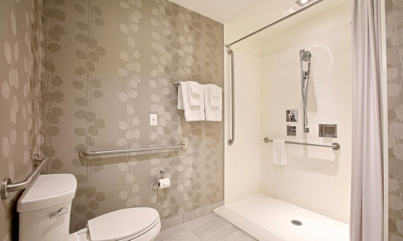 Accessible bathroom with roll-in shower, toilet, and grab bars in a tiled, beige-walled room
