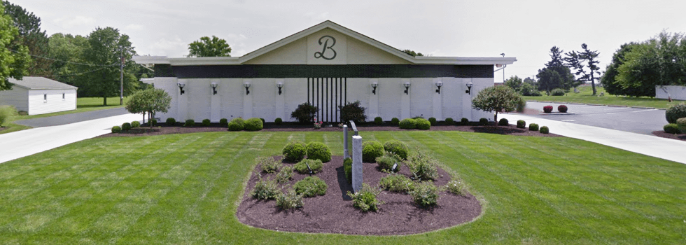 White building with gray accents, set in green grass with trees in the background.