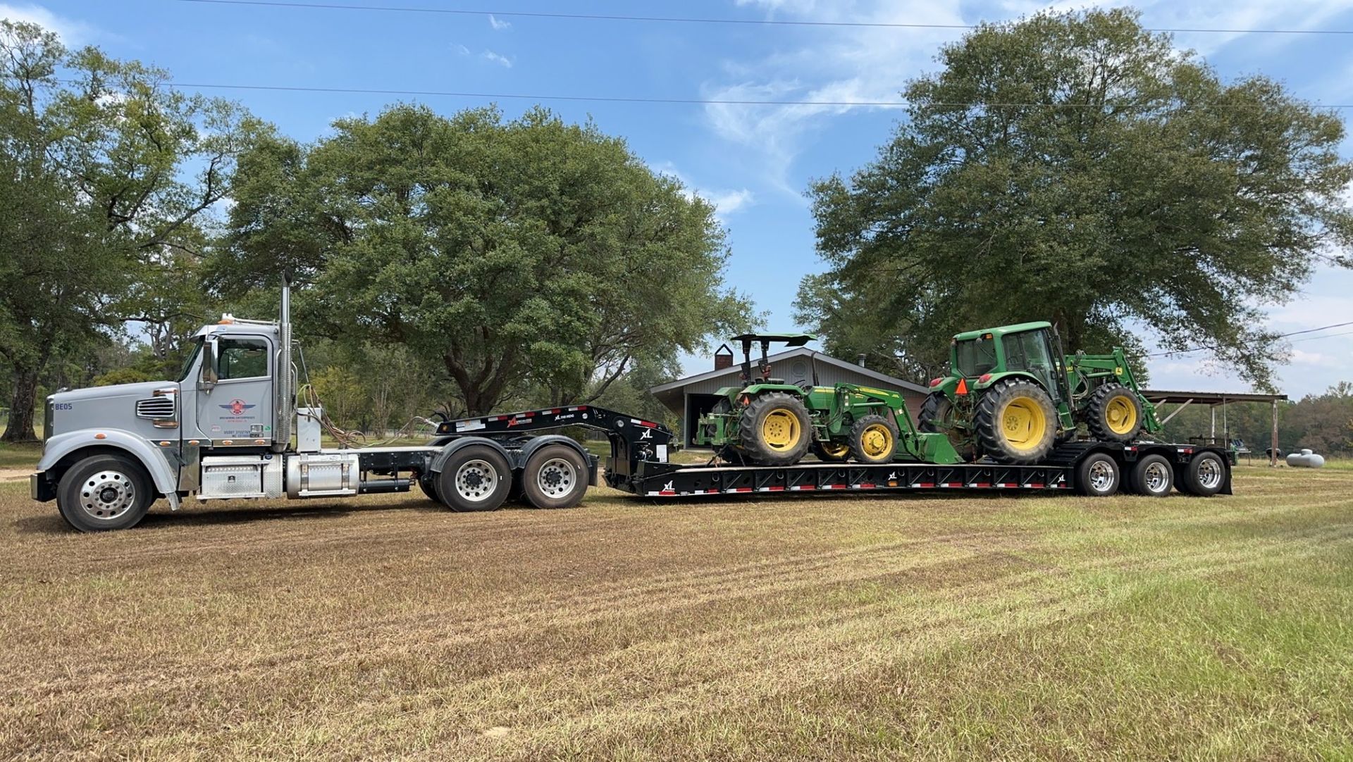 Semi-truck hauling two John Deere tractors on a trailer in a field, under a blue sky.