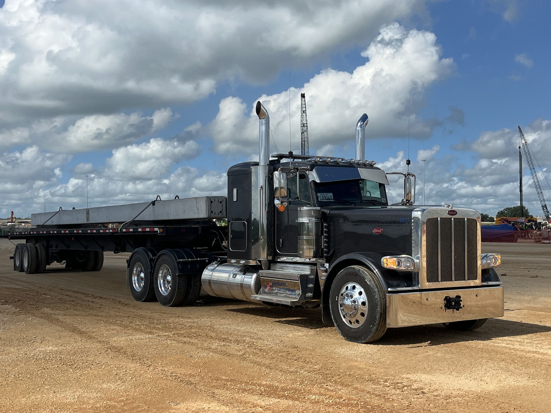 Black semi-truck with flatbed trailer on dirt, parked under a cloudy sky.