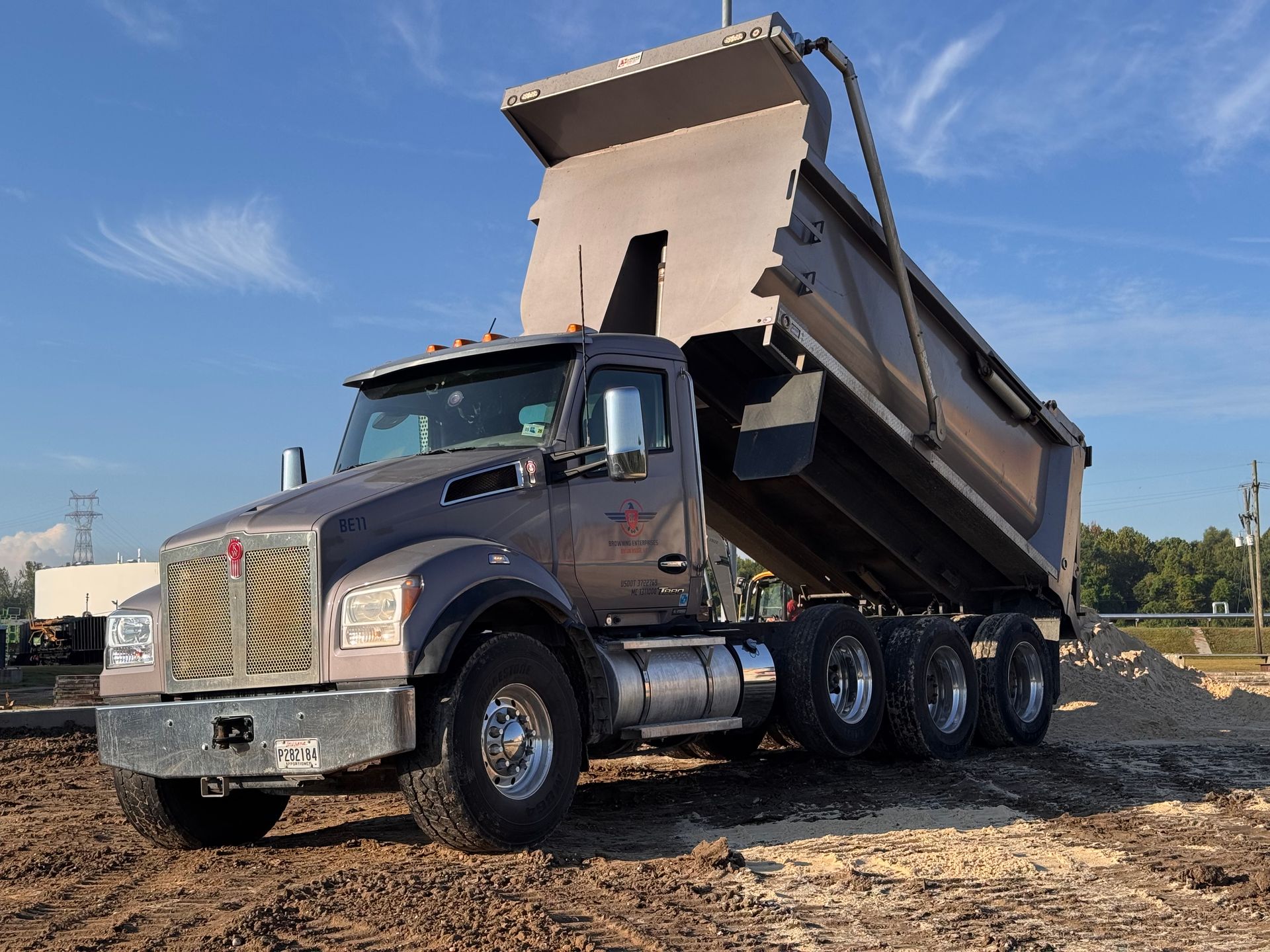 Dump truck, gray cab and bed, raised, dumping gravel at a construction site, blue sky.