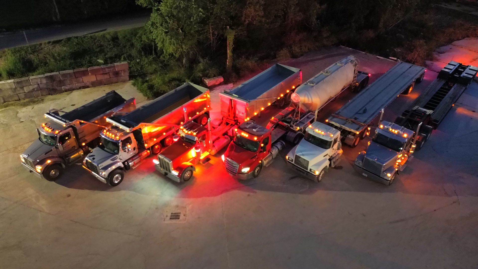 A line of semi-trucks with various trailers parked at night; lit by headlights and red/orange lights.