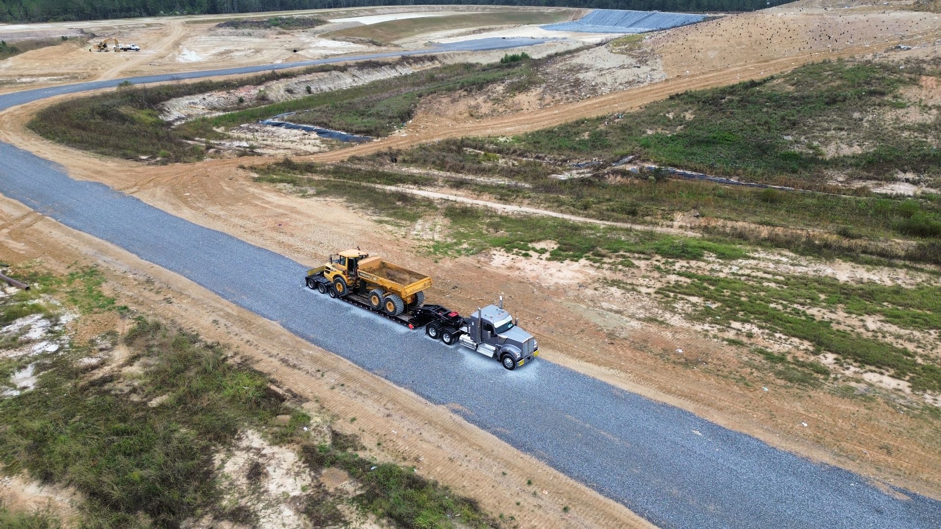 A gravel road with a truck hauling a trailer with a piece of heavy equipment. Land clearing in the background.