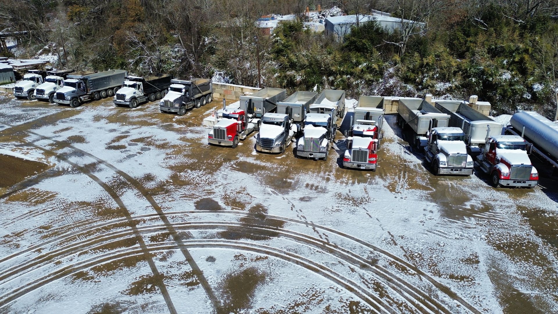 Trucks parked in a snow-covered lot. Some have red cabs. Trees and buildings are in the background.