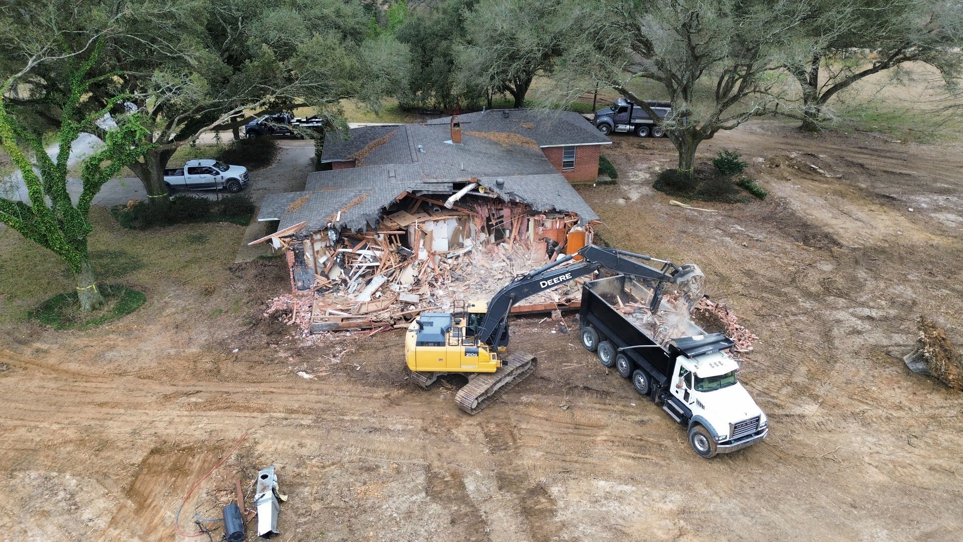 Demolition of a house, excavator and dump truck removing debris. Muddy field, trees in background.