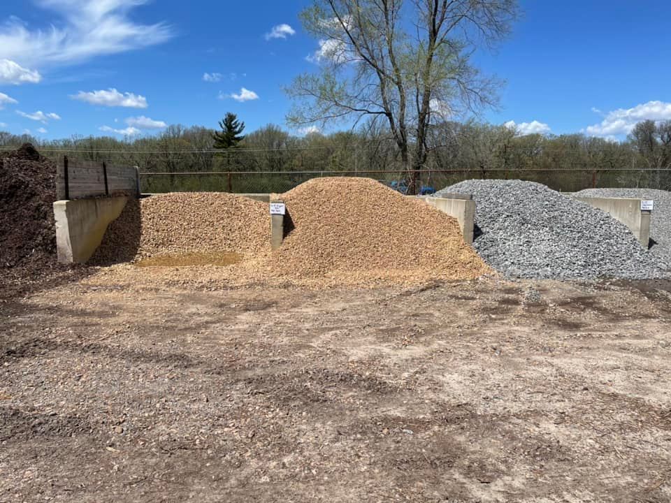 Outdoor gravel and dirt piles under a blue sky beside a dirt lot and trees