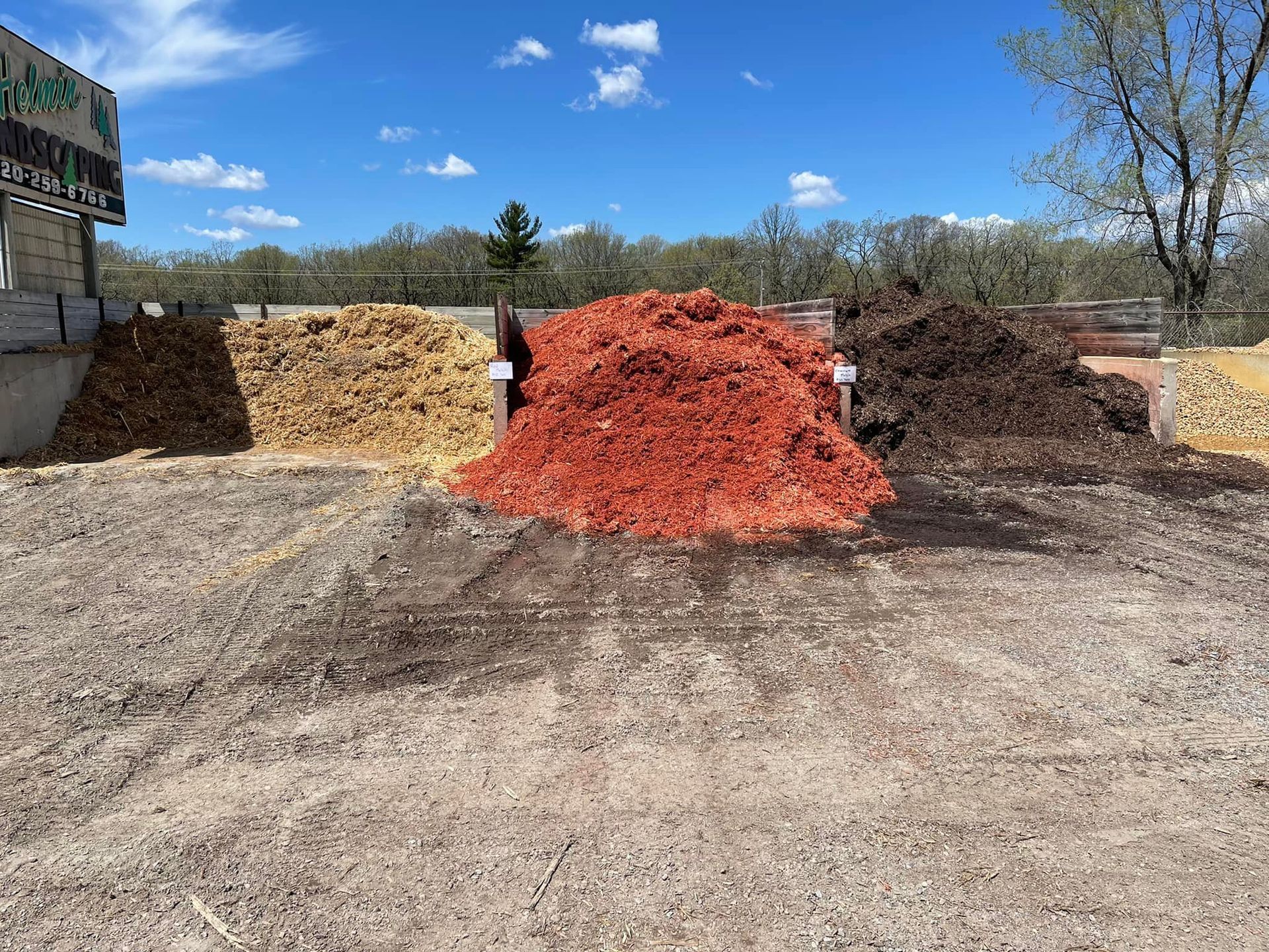 Piles of mulch in tan, red, and dark brown sit in a sunny outdoor lot under a blue sky.