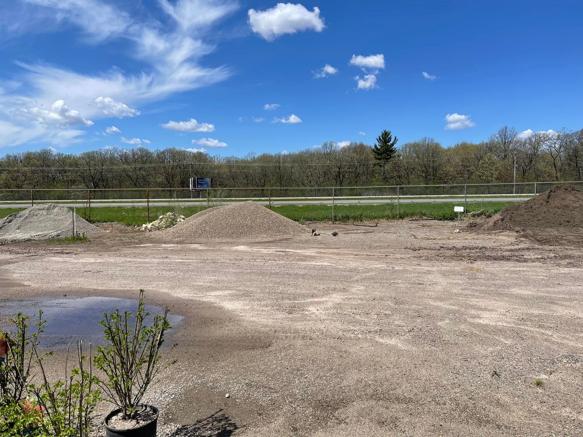 Dirt lot with gravel piles and a small puddle under a blue sky with scattered clouds