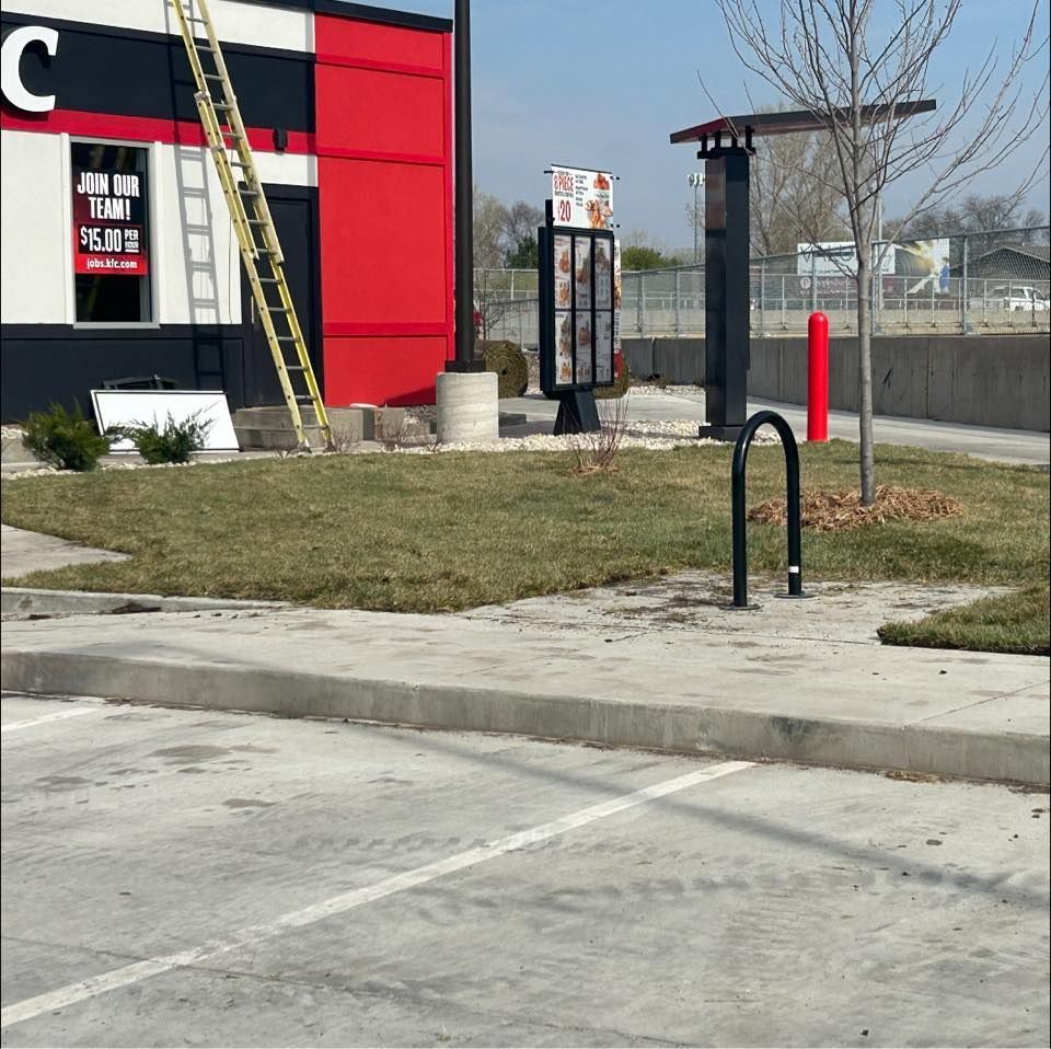 Drive-thru area by a red-and-black building with a ladder, sign, grassy strip, and curb.