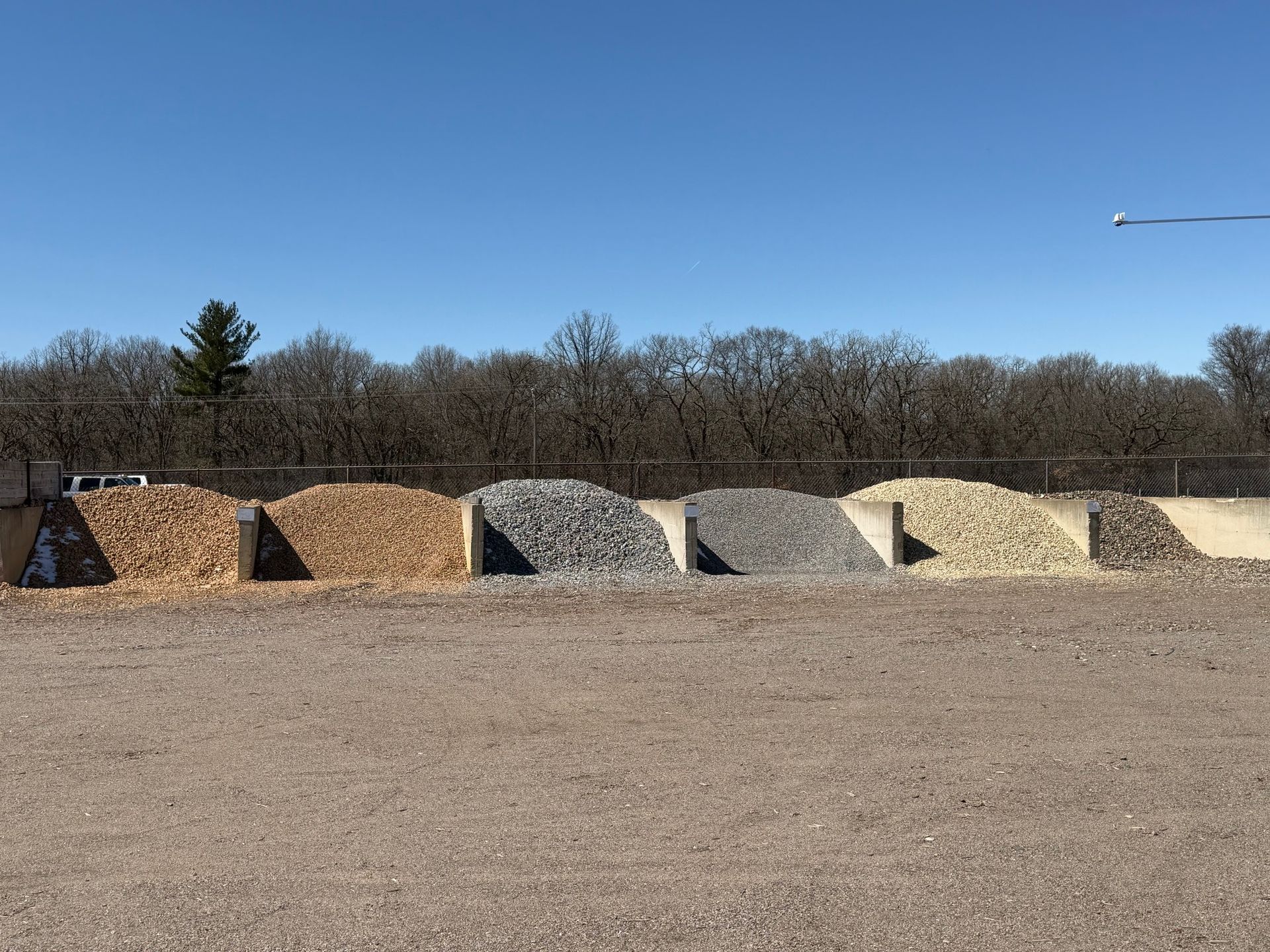 Gravel and mulch piles lined up outdoors under a clear blue sky, with trees in the background