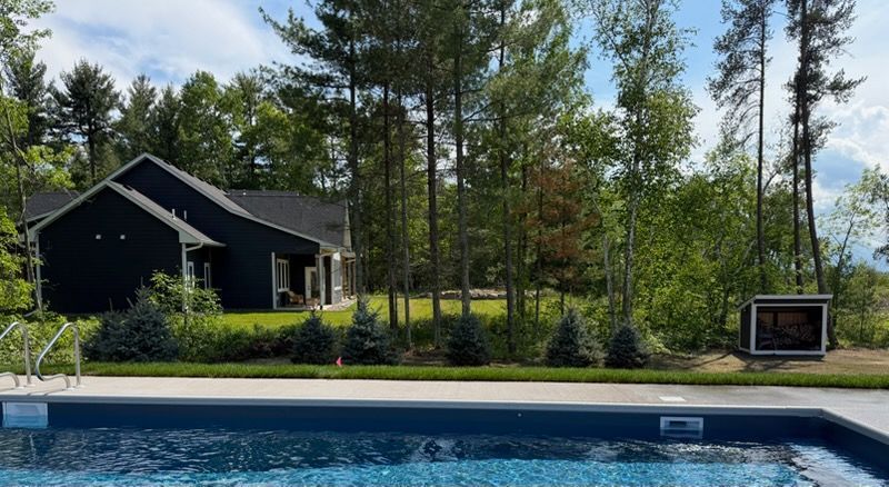 Backyard with a blue swimming pool, dark house, tall trees, and a small fire pit under a bright sky