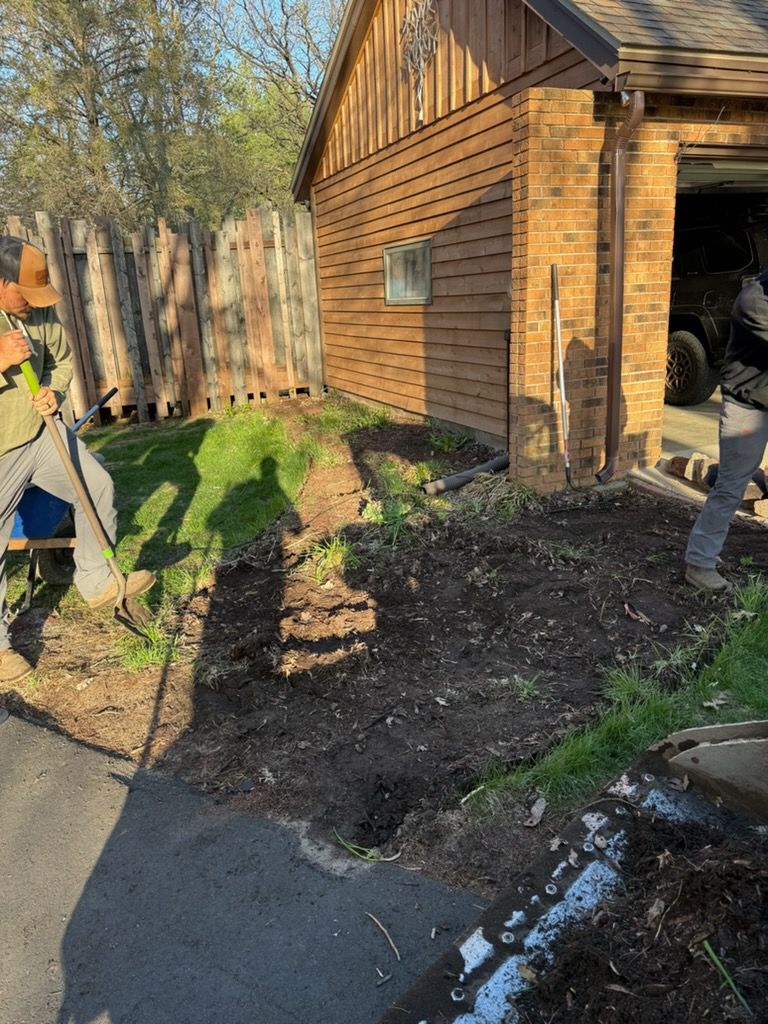 People landscaping a yard beside a brick house, with fresh mulch and a paved path.
