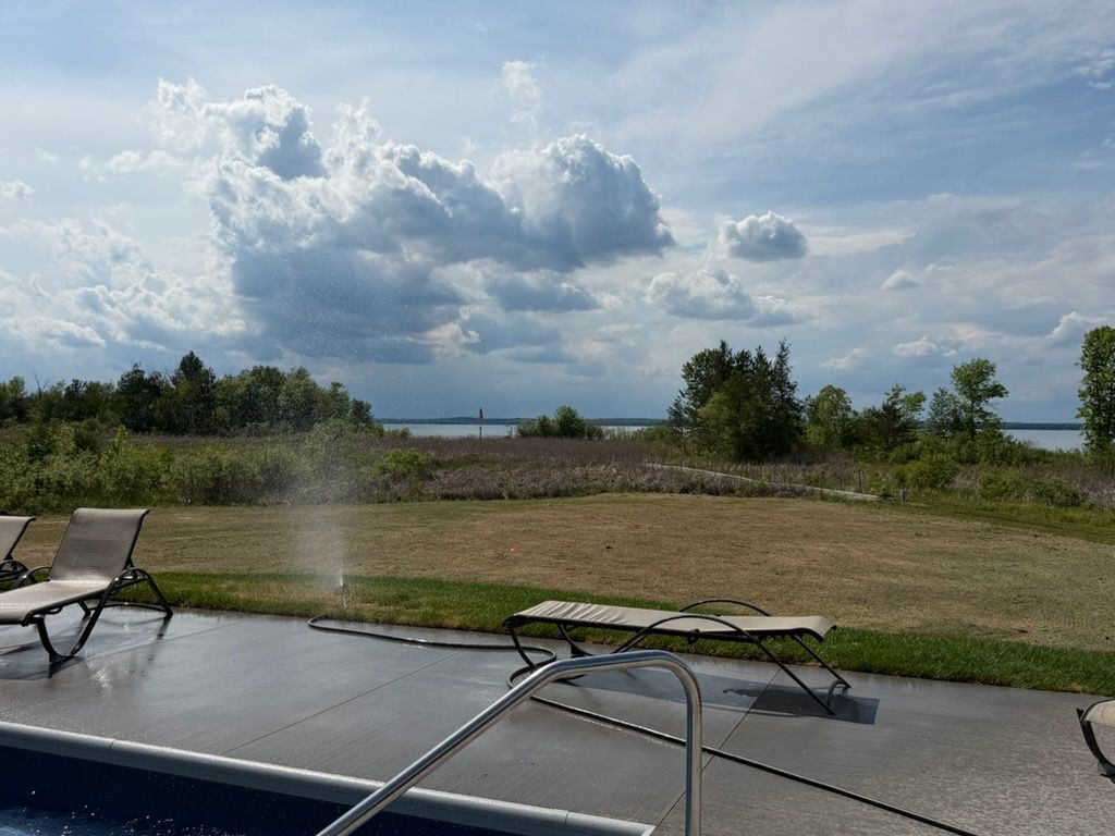Patio chairs beside a pool with a grassy field and cloudy blue sky beyond.